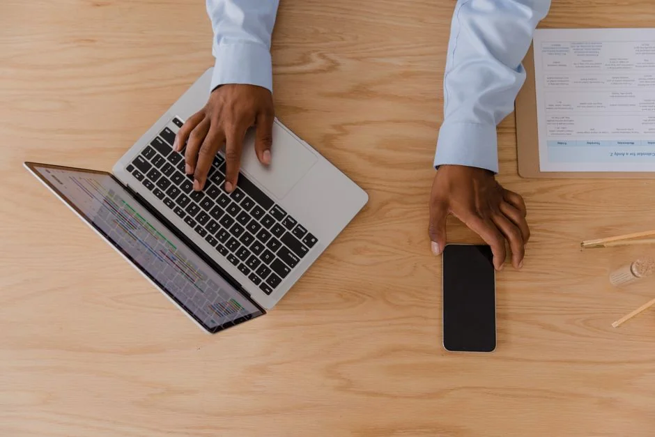 Hands holding a smartphone next to a laptop with stock charts, highlighting digital trading.